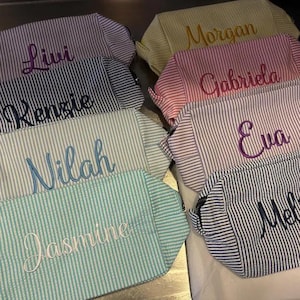 May include: A collection of rectangular zippered pouches made from seersucker fabric in various pastel and neutral striped patterns. Each pouch features a name embroidered in a cursive script, including Livi, Kenzie, Nilah, Jasmine, Morgan, Gabriela, Eva, and Mel.