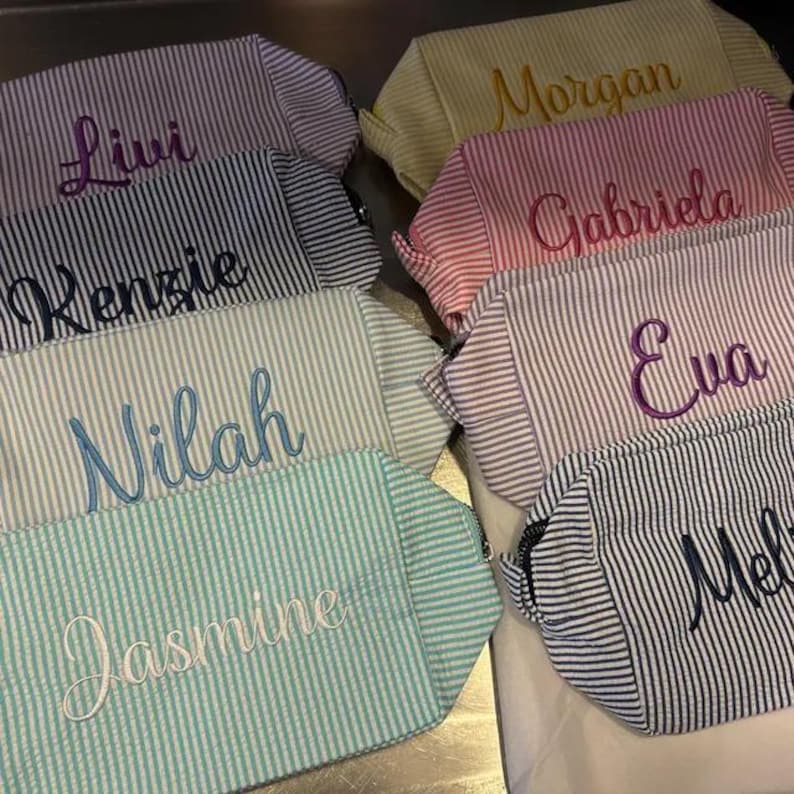 May include: A collection of rectangular zippered pouches made from seersucker fabric in various pastel and neutral striped patterns. Each pouch features a name embroidered in a cursive script, including Livi, Kenzie, Nilah, Jasmine, Morgan, Gabriela, Eva, and Mel.