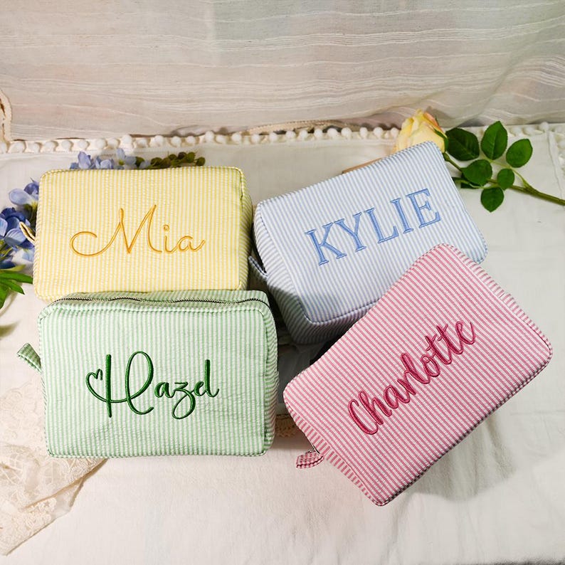 May include: Four rectangular zippered pouches in yellow, light blue, light green, and pink, each featuring a white pinstripe pattern. The pouches are embroidered with the names Mia, Kylie, Hazel, and Charlotte in matching thread colors.