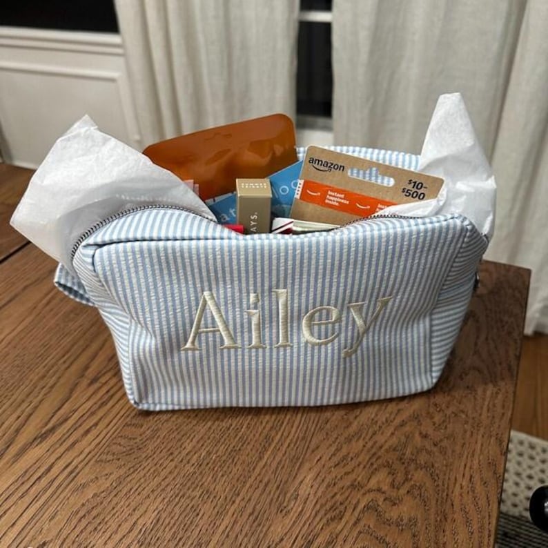 May include: A light blue and white pinstriped fabric pouch with the name Ailey embroidered in white thread on the front. The pouch is filled with white tissue paper, a brown rectangular item, and an Amazon gift card with visible text.