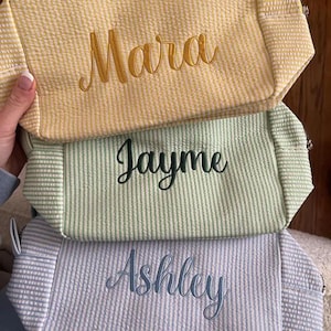 May include: Three rectangular zippered pouches made of seersucker fabric with vertical stripes. The top pouch is yellow with the name Mara embroidered in gold. The middle pouch is light green with the name Jayme in dark green. The bottom is blue with Ashley in blue.