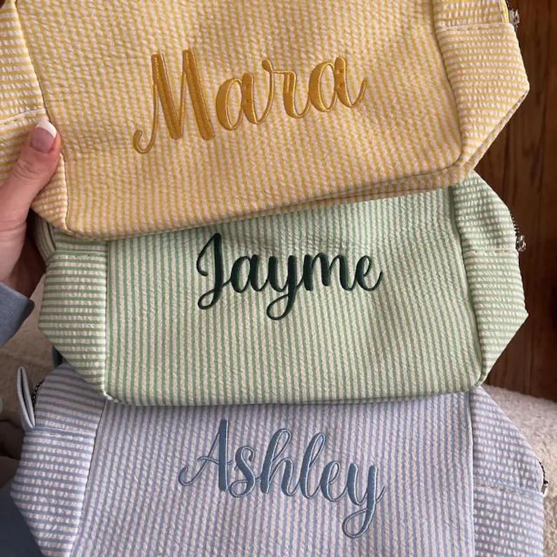 May include: Three rectangular zippered pouches made of seersucker fabric with vertical stripes. The top pouch is yellow with the name Mara embroidered in gold. The middle pouch is light green with the name Jayme in dark green. The bottom is blue with Ashley in blue.