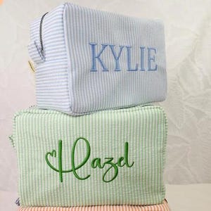 May include: Two rectangular zippered pouches made of seersucker fabric with thin vertical stripes. The top pouch is light blue with the name KYLIE embroidered in blue. The bottom pouch is light green with the name Hazel embroidered in green.