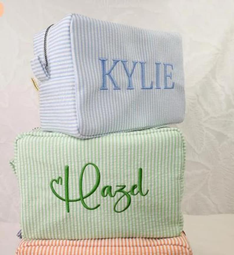 May include: Two rectangular zippered pouches made of seersucker fabric with thin vertical stripes. The top pouch is light blue with the name KYLIE embroidered in blue. The bottom pouch is light green with the name Hazel embroidered in green.