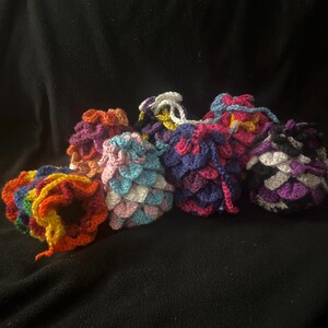 May include: A collection of handmade crochet pouches in various colors, including rainbow, orange, pink, blue, purple, and black. Each pouch features a textured, scale-like pattern and a drawstring closure, set against a black background.