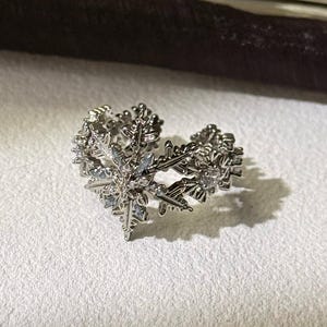 Puede incluir: Un anillo plateado en forma de corazón, con un diseño de copo de nieve. El anillo está sobre una superficie blanca, con un fondo marrón oscuro y dorado. Los intrincados detalles del anillo se destacan por la luz.