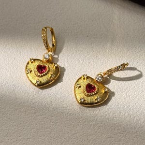 May include: A pair of gold heart-shaped earrings. Each earring has a red heart-shaped stone in the centre, surrounded by small clear stones and a textured gold border. The earrings have a hinged closure.