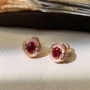 May include: A pair of stud earrings featuring oval-cut red gemstones set in a gold frame. The earrings have a white, oval-shaped border and are accented with small, sparkling stones. The design is elegant and suitable for formal occasions.