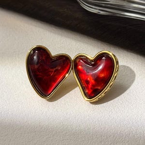 Może przedstawiać: Para złotych kolczyków w kształcie serca z czerwonymi, marmurowymi wstawkami ze szkła.