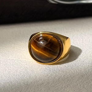 May include: A gold ring featuring a large, round tiger's eye gemstone. The stone displays brown and gold hues, set within a simple, rectangular gold band. This ring is a bold accessory, suitable for daily wear.
