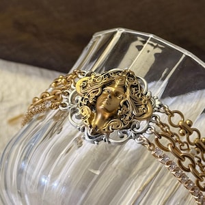 Puede incluir: Una pulsera dorada y plateada con un camafeo de rostro detallado. La pulsera tiene múltiples hilos de cadena dorada y un engaste de filigrana plateada alrededor del camafeo. La pulsera se exhibe sobre una superficie de vidrio transparente.