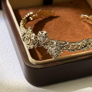 May include: A gold-toned metal cuff bracelet featuring a textured, organic surface that resembles tree bark. The bracelet is adorned with three detailed, raised rose flower motifs along the front, showcasing a polished, reflective finish.