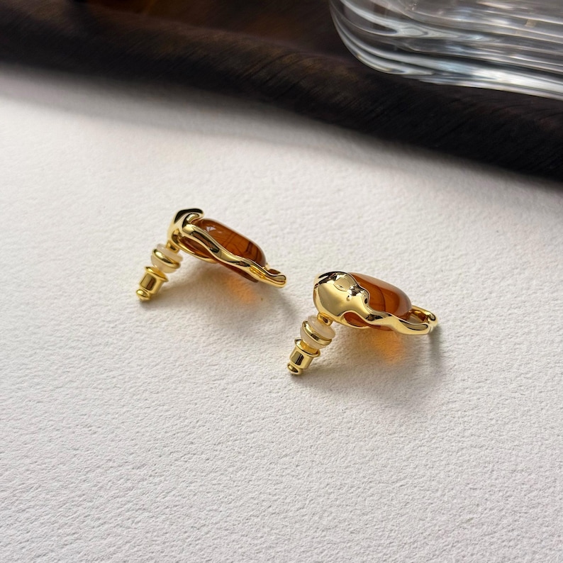 May include: A pair of gold earrings with amber-colored stones. The stones are set in a gold frame with a wavy design.