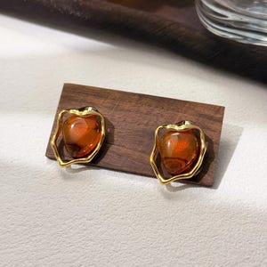 May include: A pair of gold-toned earrings with a brown, amber-like stone set in an irregular, organic-shaped frame.