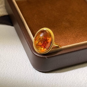 Op de afbeelding: Een gouden ring met een grote, ovale barnsteen. De barnsteen vertoont een mix van oranje en roodbruine tinten, met zichtbare insluitsels. De ring heeft een gedetailleerde, getextureerde gouden zetting en een eenvoudige band.