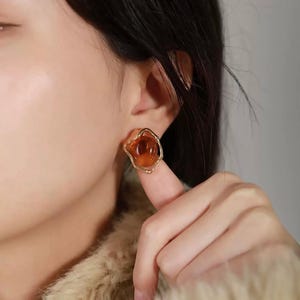 May include: A pair of gold-toned earrings with a brown resin flower design. The earrings are small and delicate.
