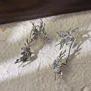 May include: A pair of silver-toned ear cuffs and a single earring, each featuring a detailed butterfly wing design. The jewelry is displayed on a textured, off-white surface, with a dark brown background.