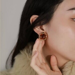 May include: Gold-tone clip-on earrings with a brown, oval-shaped stone set in a textured, flower-shaped frame.