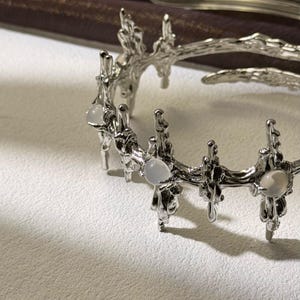 May include: A silver-toned bracelet with an organic, branch-like design. The bracelet features several round, pale gemstones set along its length. The intricate metalwork has a textured, almost rough appearance, creating a unique jewelry piece.