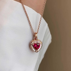 May include: A rose gold necklace featuring a heart-shaped pendant. The pendant has a vibrant red gemstone center, surrounded by a white border and accented with small, sparkling crystals. The chain is delicate and complements the pendant's design.