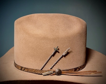 May include: A tan felt cowboy hat with a snakeskin band and a silver cross and feather concho. The hat has a leather hatband with a tassel.