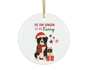 May include: A white ceramic Christmas ornament with a gold string. The ornament features a black and white dog wearing a Santa hat and scarf, with gifts and stars. Text reads "Tis the season to be Furry."
