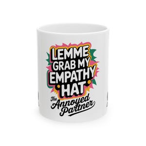 May include: White ceramic mug with the text "LEMME GRAB MY EMPATHY HAT" in a colorful design. The phrase "The Annoyed Partner" is written below in a script font. The mug is a standard size.