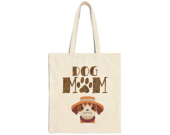 May include: Cream-colored tote bag with the words "DOG MOM" in a leopard print font. A cartoon dog wearing a hat is featured below the text. The bag has two handles and is made of a canvas-like material.