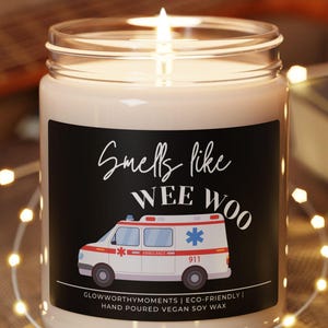 May include: A lit candle in a glass jar with a black label featuring an ambulance graphic and the text "Smells like WEE WOO". The candle sits on a wooden coaster, surrounded by fairy lights.