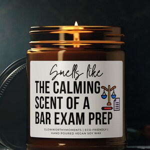 Puede incluir: Una vela encendida en un frasco de vidrio marrón con tapa negra. La etiqueta dice "Smells like the calming scent of a bar exam prep". El frasco está sobre una superficie oscura, con el texto "Glowworthy Moments | Eco-friendly | Hand poured vegan soy wax."