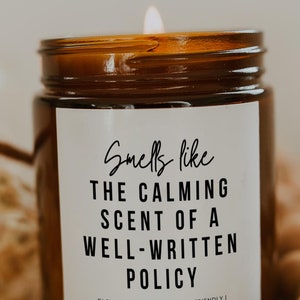 May include: A brown glass candle jar with a white label that reads "Smells like THE CALMING SCENT OF A WELL-WRITTEN POLICY" and "GLOWWORTHYMOMENTS | ECO-FRIENDLY | HAND POURED VEGAN SOY WAX". The candle is lit and the flame is visible.