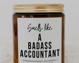 May include: A brown glass candle jar with a gold lid. The label reads "Smells like a badass accountant" and includes the brand name "Glowworthy Moments" and the text "Eco-friendly | Hand poured vegan soy wax".