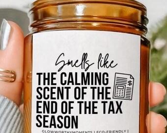 May include: A brown glass candle with a white label that reads "Smells like the calming scent of the end of the tax season". The label also includes a small black and white illustration of a calculator with a dollar sign.