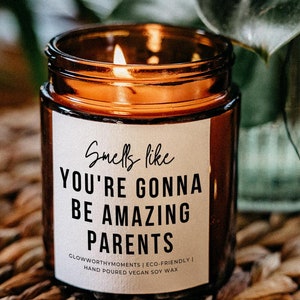 May include: A brown glass candle with a white label that reads "Smells like You're Gonna Be Amazing Parents". The candle is lit and the flame is visible. The candle is sitting on a woven basket.
