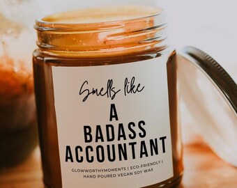 May include: A brown glass candle jar with a white label that reads "Smells like a Badass Accountant". The label also includes the text "Glowworthy Moments | Eco-Friendly | Hand Poured Vegan Soy Wax".