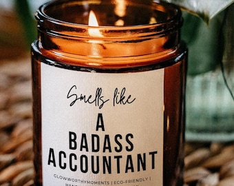 May include: A brown glass candle with a white label that reads "Smells like a badass accountant". The label also includes the text "Glowworthy Moments | Eco-Friendly | Hand Poured Vegan Soy Wax".