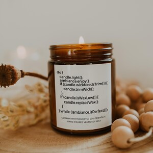 Op de afbeelding: Een bruine glazen kaars met een wit label waarop staat "do { candle.light(); ambiance.enjoy(); if (candle.wickNeedsTrim()) { candle.trimWick(); } if (candle.isWaxLow()) { candle.replaceWax(); } } while (!ambiance.isPerfect()); GLOWWORTHYMOMENTS | ECO-FRIENDLY | HAND POURED VEGAN SOY WAX". De kaars is aangestoken en de vlam is zichtbaar.