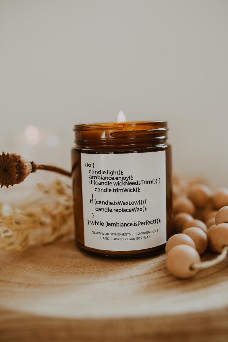 Op de afbeelding: Een bruine glazen kaars met een wit label waarop staat "do { candle.light(); ambiance.enjoy(); if (candle.wickNeedsTrim()) { candle.trimWick(); } if (candle.isWaxLow()) { candle.replaceWax(); } } while (!ambiance.isPerfect()); GLOWWORTHYMOMENTS | ECO-FRIENDLY | HAND POURED VEGAN SOY WAX". De kaars is aangestoken en de vlam is zichtbaar.