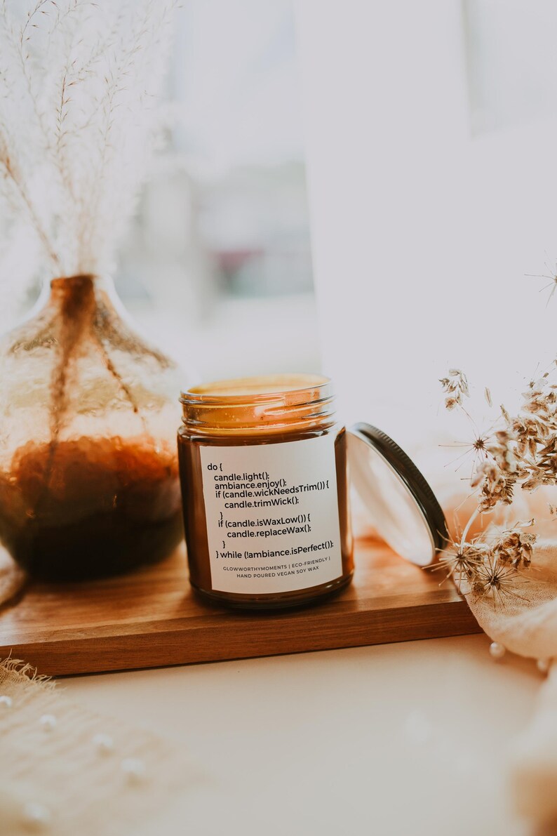 Op de afbeelding: Een bruine glazen kaars pot met een wit label met de tekst "do ( candle.light() ambiance.enjoy() if (candle.wickNeedsTrim()) { candle.trimWick() if (candle.isWaxLow()) { candle.replaceWax() } while (ambiance.isPerfect()) } GLOWWORTHYMOMENTS | ECO-FRIENDLY | HAND POURED VEGAN SOY WAX"