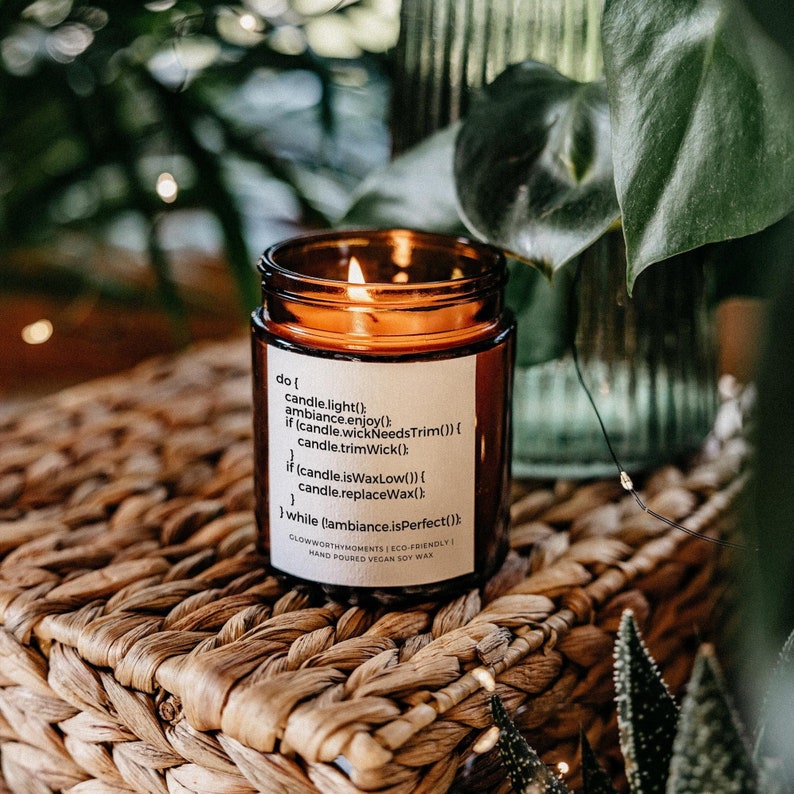 Op de afbeelding: Een bruine glazen kaars pot met een wit label waarop staat "do { candle.light(); ambiance.enjoy(); if (candle.wickNeedsTrim()) { candle.trimWick(); } if (candle.isWaxLow()) { candle.replaceWax(); } while (ambiance.isPerfect()); } GLOWWORTHYMOMENTS ECO-FRIENDLY HAND POURED VEGAN SOY WAX". De kaars is aangestoken en de vlam is zichtbaar.