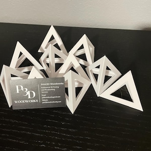 May include: A group of white triangular geometric shapes arranged on a black surface. A business card with the text "Print3D Woodworks" is in the foreground.