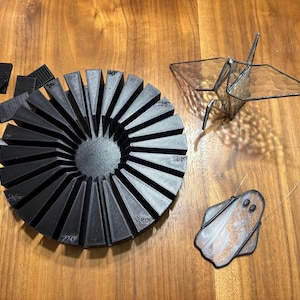 May include: A collection of handmade items on a wooden surface. Includes a black, fan-shaped object with degree markings, a glass origami crane, and a ghost-shaped ornament. Additional black geometric shapes are also present.