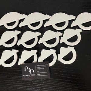 May include: A set of ten white plastic templates for woodworking projects. Each template is shaped like a half-circle with a different size marked on it. The templates are arranged in a circle on a black surface. The text "Print2D Woodworks" is visible on a business card in the lower right corner of the image.