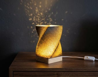 May include: A modern, sculptural table lamp with a twisted design. The lamp has a textured, dark brown exterior and emits a warm, golden light. It sits on a light-colored wooden base and includes a white power cord with an on/off switch.