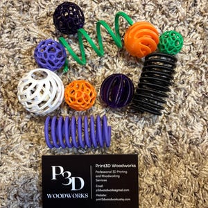 May include: A collection of 3D-printed objects in various colors, including spheres, spirals, and a coil. Colors include purple, orange, green, white, and black. A business card for Print3D Woodworks is also present.