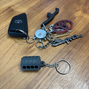 May include: A collection of car keys and keychains on a wooden surface. Includes a black Toyota key fob, a black wrench-shaped keychain, a red and black Toyota logo keychain, and a silver "Supra" keychain. A gray engine block keychain is also present.