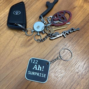Op de afbeelding: Een verzameling sleutels en sleutelhangers op een houten oppervlak. Inclusief een zwarte Toyota-sleutelhanger, een zwarte flesopener, een rood-zwarte Toyota-logo sleutelhanger en een zilveren Supra sleutelhanger. Een vierkante sleutelhanger met de tekst "122 Ah! SURPRISE" is ook aanwezig.