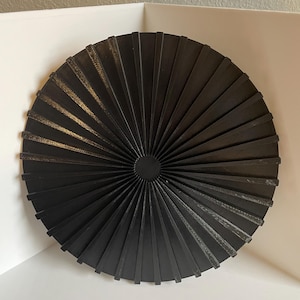 May include: A black circular wall decor with a fan-like design. The decor is made of multiple thin, black strips that radiate outwards from the center.