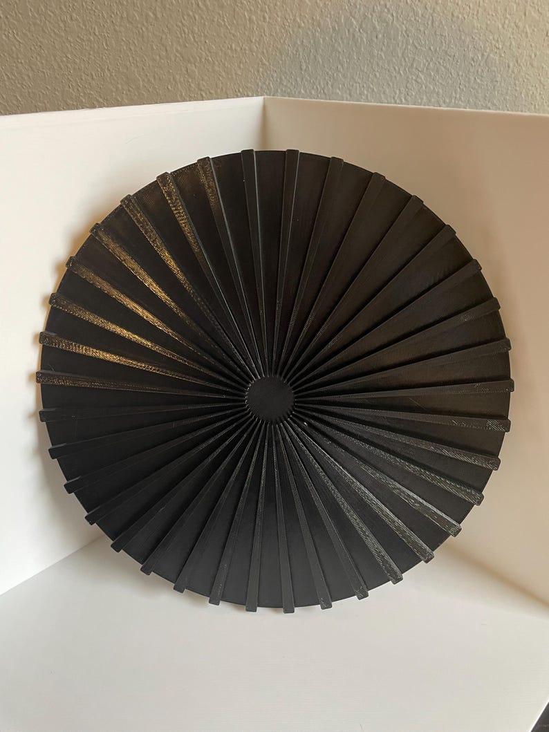 May include: A black circular wall decor with a fan-like design. The decor is made of multiple thin, black strips that radiate outwards from the center.