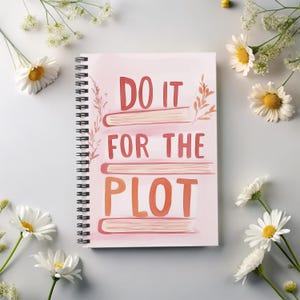 May include: A pink spiral notebook with the text "DO IT FOR THE PLOT" in orange and pink lettering. The notebook is surrounded by white flowers.